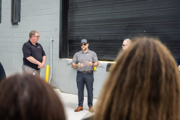 Three men in polo shirts stand outside a gray industrial building while a speaker in sunglasses talks to a small audience partially visible in the foreground.