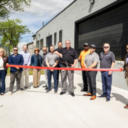 Group of people posing with a red ribbon in front of a gray industrial building, suggesting a ribbon-cutting ceremony.