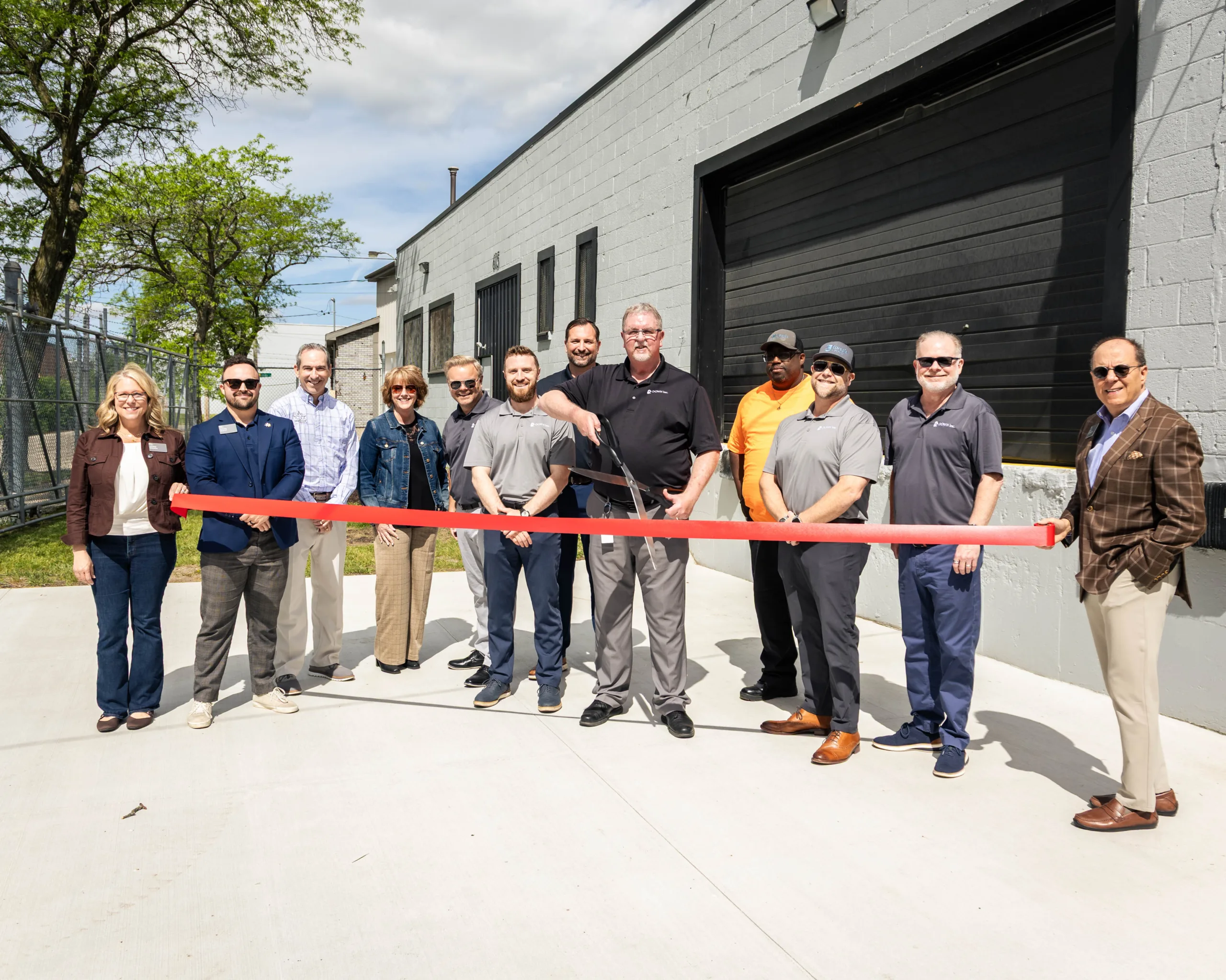 Group of people posing with a red ribbon in front of a gray industrial building, suggesting a ribbon-cutting ceremony.