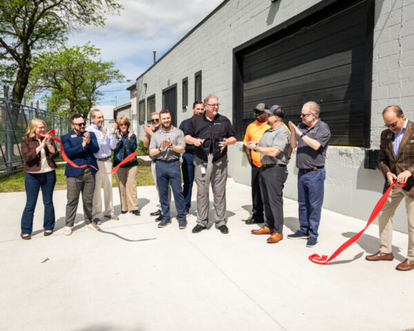 Group of professionals outdoors at a ribbon-cutting celebration beside a gray industrial building.