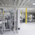 Industrial factory floor with fenced enclosures housing machinery and yellow safety posts under a white ceiling.